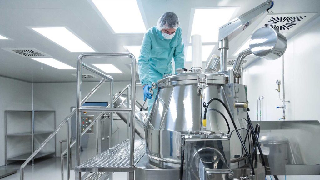 Lab technician in scrubs, hairnet, and mask adjusts a large stainless steel bioprocess vessel in a cleanroom.